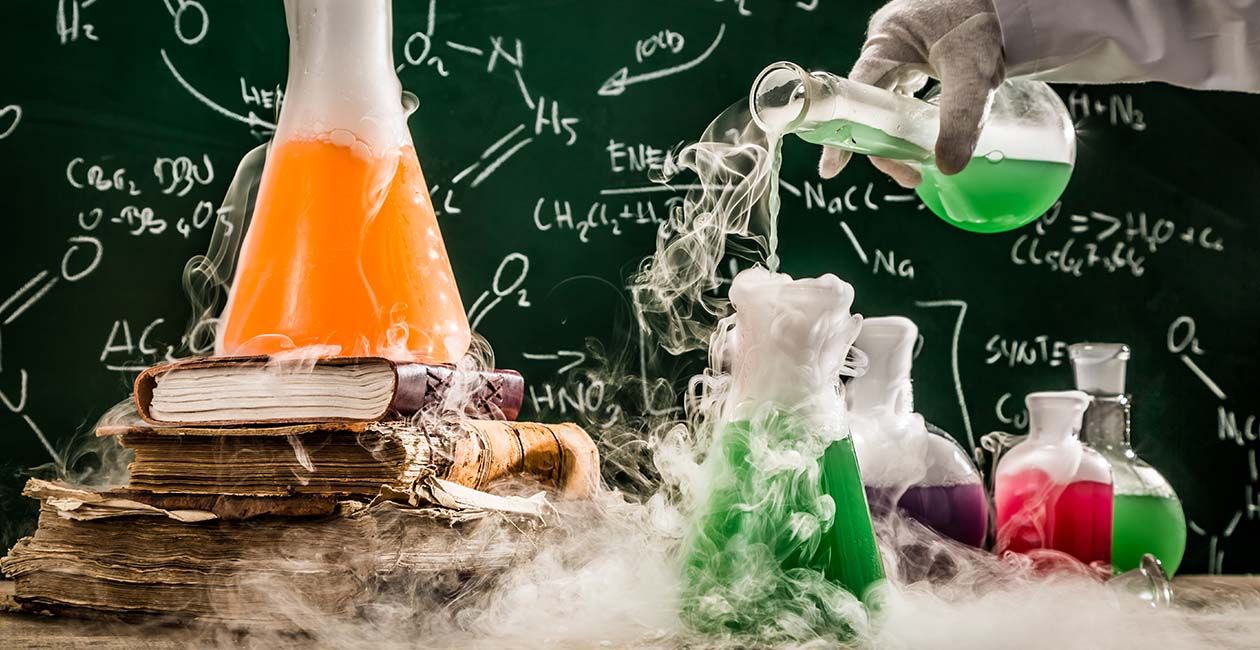 Hand pouring liquid into beaker on a desk.  Another beaker on a stack of books with smoke surrounding all beakers and table