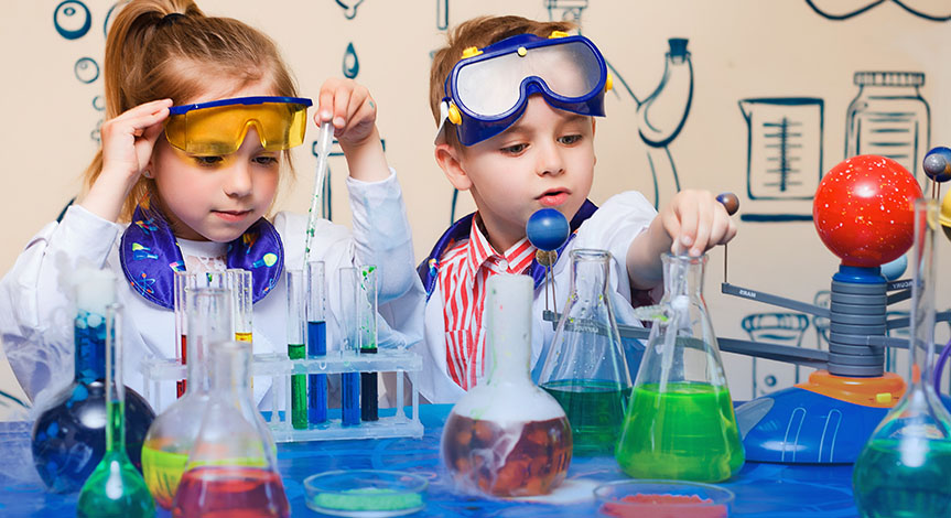 A boy and girl taking liquid from beaker using a pipette. On the table there are beakers and test tubes with colored liquids
