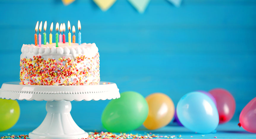 Delicious birthday cake on a white tray with burning candles and colorful balloons on table