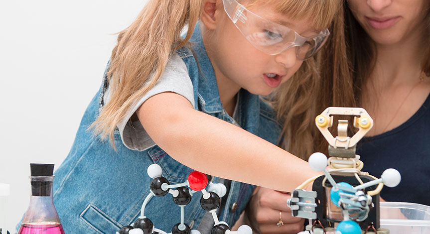 A woman teacher and girl collect molecules and conduct experiments. On the table are a robot and beakers with liquid inside.