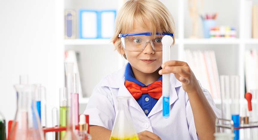 Boy holding a test tube filled with yellow liquid.  He is surrounded by other beakers and test tubes with multi colored liqui